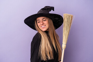 Wall Mural - Young Russian woman disguised as a witch holding a broom isolated on purple background looks aside smiling, cheerful and pleasant.