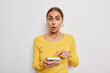 © wayhome.studio  - Isolated shot of surprised young Caucasian woman stares surprised indicates at modern smartphone screen cannot believe in shocking amazing news wears yellow sweater isolated over white background.