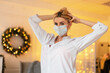 Pretty young girl with a protective medical mask in a white shirt is doing her hair on the background of Christmas decorations and yellow lights in the room. COVID-19 concept and female beauty