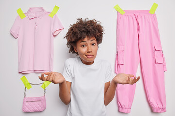 Wall Mural - Clueless doubtful young woman with Afro hair spreads palms feels hesitant shrugs shoulders wears white t shirt poses against t shirt trousers and bag plastered to white background. So what to do