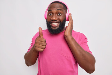 Wall Mural - Positive dark skinned man with thick beard smiles broadly shows excellent sign keeps thumb up listens music in wireless headphones wears pink t shirt isolated over white background. I like this