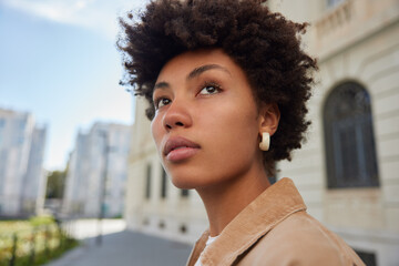 Wall Mural - Photo of good looking young woman with Afro hair dressed in stylish clothes looks into distance thinks about something while returning from excursion stands outdoor against blurred background.