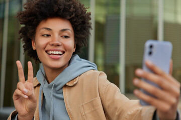 Wall Mural - attractive positive african american girl holds mobile phone makes peace gesture takes selfie walks 
