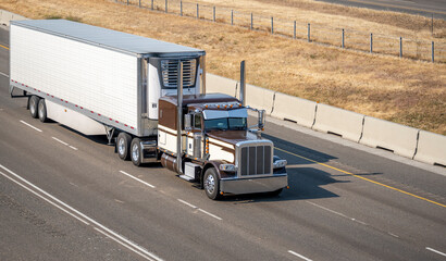 Brown classic American idol big rig semi truck with vertical exhaust pipes transporting frozen cargo in reefer semi trailer running on the summer highway road