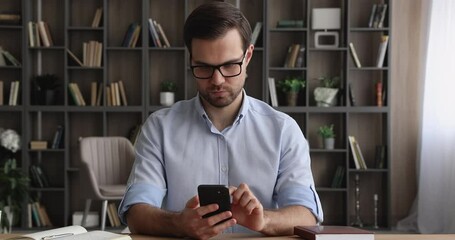 Canvas Print - Happy young male entrepreneur in eyeglasses sitting at table using mobile software application, texting message communicating in social network, responding mobile email, business telecommute concept.