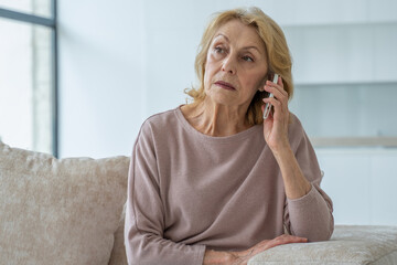 a sad elderly woman is talking on a mobile phone at home sitting on the couch