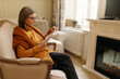 © shurkin_son - Woman of 60s in living room reading book in front of electric fireplace in cozy stylish chair, wearing glasses, jeans and orange blouse with cardigan over shoulders. People and lifestyle