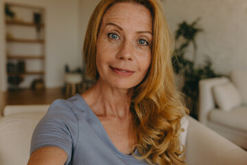 Wall Mural - Selfie of middle-aged ginger head long-haired woman doing selfie at home, posing with happy, cheerful face expression and sincere look, wearing blue clothes, having some facial lines and freckles