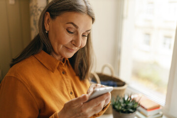 Stylish cheerful middle aged woman posing at window, holding smart phone, typing text message, communicating with friends via group online chat in messenger application. Retired lady texting sms