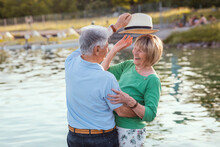 Senior Man Putting Hat On Cheerful Woman By Lake