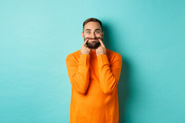 Image of bearded man stretching lips in happy smile, faking happiness, standing over light blue background