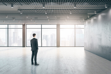 Poster - Attractive thoughtful young european businessman standing in empty interior with windows, city view and daylight.