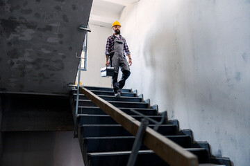 Wall Mural - A bearded handyman with a helmet on his head with a toolbox in his hands is going down the stairs in a new building in the building process. There is a lot of work to do.