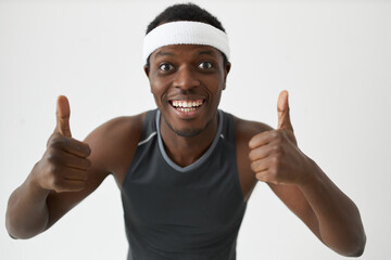 Wall Mural - Joyful youthful Afro male in fitness clothes and headband smiling showing white healthy teeth and putting thumbs up, expressing feelings of excellent mood and satisfaction with perfect sports results