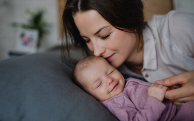 happy young mother kissing her newborn baby girl, lying on sofa indoors at home.