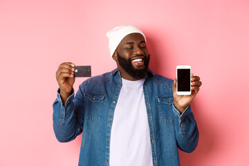 Online shopping. Satisfied Black man nod in approval, smiling and looking at phone, showing credit card and smartphone screen, standing over pink background