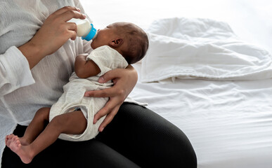 Wall Mural - Mother Sitting on white bed, and feed milk from bottle milk to her 12-day-old baby black skin newborn son, to family and food for infant concept.