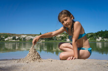 A Little Cute Girl In A Swimsuit Is Building A Sand Castle On The Shore Of A Blue Quarry Lake.