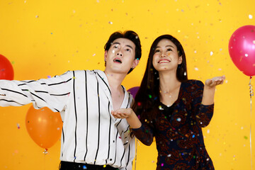 Studio shot of Asian happy young male and female lover couple model in fashionable wears playing fun with shiny paper confetti in celebration party with balloons on yellow background