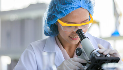 Wall Mural - Close up shot of Asian female professional scientist in white lab coat safety glasses hygiene cap and rubber gloves using scientific microscope lens looking zooming sample on laboratory working desk