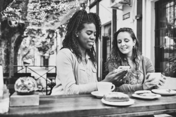 Two nice young woman watch mobile phone, drink coffee and eat cakes