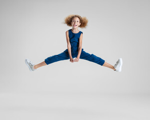 Wall Mural - One little Caucasian cute girl in blue stylish jumpsuit jumping high and having fun isolated over white studio background.