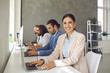 © Studio Romantic - Business people using modern computers in office. Portrait of happy cheerful beautiful young woman sitting at table with team of colleagues, working on project on laptop, looking at camera and smiling