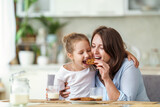 Fototapeta Łazienka - happy loving family, mom and daughter, playing sitting at table and having breakfast at home in morning. woman and girl eat oatmeal cookies and drink cow's milk, and have nice time together in kitchen