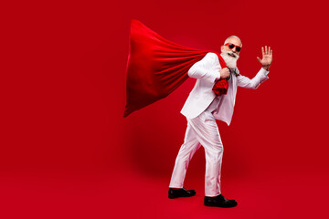 Wall Mural - Photo of carefree cute man dressed white christmas costume dark glasses holding big sack waving arm isolated red color background