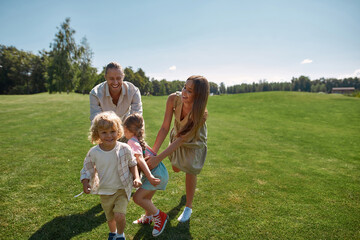 Joyful parents with two little kids playing catch together in green park on a summer day. Happy family enjoying leisure activity