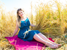 A Blonde Woman In A Turquoise Dress Sits On A Magenta Bedspread Against A Background Of Golden Ears Of Wheat In Field. Picnic On A Sunny Autumn Day In Nature