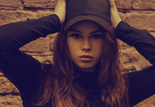 Young Sexy Young Woman Looking Success Near The Red Brick Wall On The Street Background In Black Style Clothing And Cool Cap. Autumn Season Clothing. Closeup