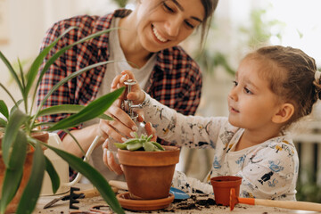 Wall Mural - Young happy mixed race woman with her little daughter is planting and watering houseplants at home.Home gardening.Family leisure, hobby concept.Biophilia design and urban jungle concept.