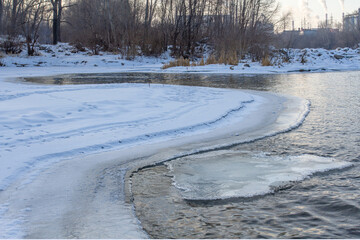 Winter landscape with a partially frozen river. Ice floats in open water. In the background there is a forest and industrial facilities.