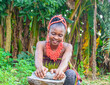 A pretty African lady or woman with beads on her head is grinding something with local grinding stone in a banana farm
