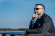 © Людмила Чиговская - A young man in a black coat and sunglasses stands on an old wooden bridge and looks into the distance. A young, handsome businessman on the riverbank.