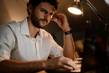 Brunette man wearing shirt working with laptop while sitting at desk