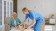 © AboutLife - Positive young nurse carries tray with breakfast to senior woman patient sitting in light living room