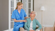 © AboutLife - Cheerful nurse in uniform listens to senior patient while sitting on armrest of comfortable armchair in room