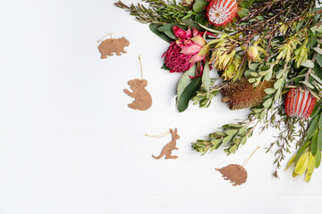 Beautiful Christmas flat lay floral arrangement of mostly Australian native flowers, Purple and Red Banksia, Red Waratah and green foliage, decorated by native wooden animals, on a white background. 