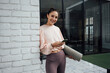 © Westend61 - Smiling woman with mobile phone and exercise mat leaning on brick wall