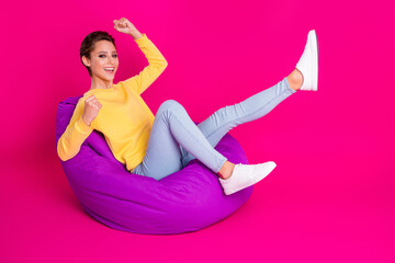 Poster - Photo of charming lucky young lady wear yellow jumper smiling sitting bean bag rising fists isolated pink color background