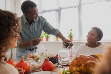 Wall Mural - Mature african american man pouring wine near family during thanksgiving celebration