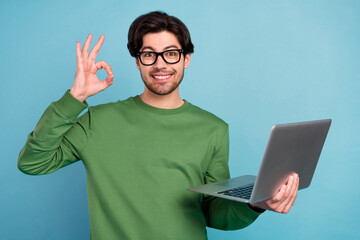 Sticker - Portrait of smiling geek nerd young businessman in glasses show okay sign hold laptop isolated on blue color background