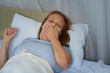 © evafesenuk - Senior woman in blue pajamas yawns while lying in bed, covering her mouth with hand