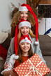 © ako-photography - three happy young women in red dwarf hats sitting on sofa and holding Chistmas gifts presents boxes at home living room