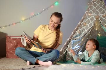 Wall Mural - Young woman reading fairy-tale to her little daughter late in evening at home