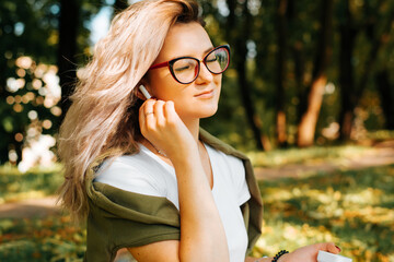 Portrait of an attractive young woman in glasses uses wireless earphones, listens music, smiling beautiful girl with closed eyes enjoys sound in headphones outdoors in park. People and technology