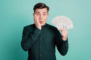 Portrait of surprised funny millionaire guy hold fan cash palm cheek wow reaction on turquoise background