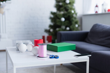 Canvas Print - table with gift box, bobbin with decorative ribbon, baubles and scissors in living room with blurred christmas tree